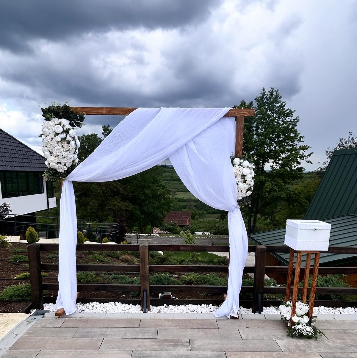 Drveni venčani luk sa belom draperijom i cvetnim aranžmanom na otvorenom